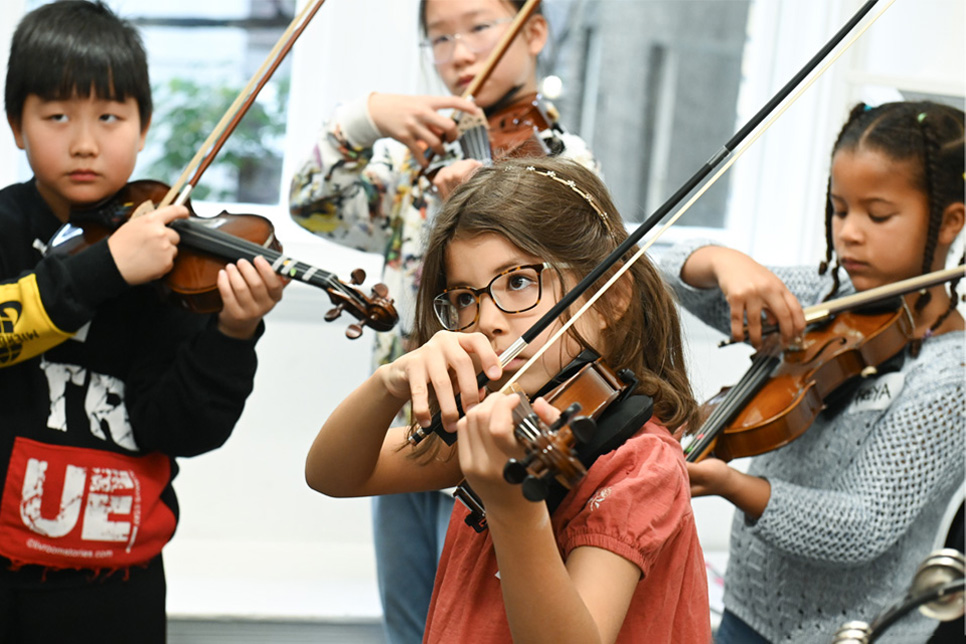 A group of young children, wearing normal clothes, all playing the violin in a group teaching session.